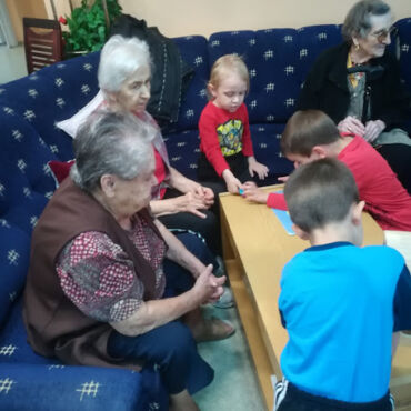 Az otthon lakói figyelik, ahogy a gyermekek játszanak az asztal körül.